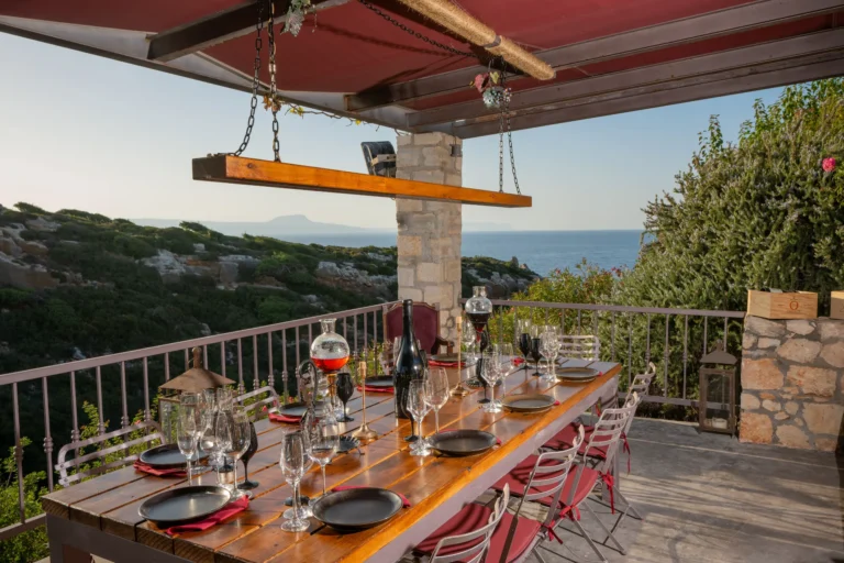 Outdoor balcony with gorge and sea view, set up with a table, chairs, utensils, and wine glasses for wine tasting.